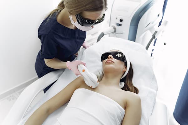 woman laying on clinic bed with another woman applying a laser hair removal treatment
